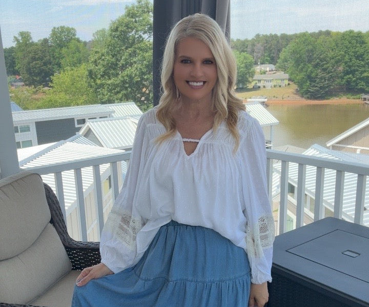 Woman in a white blouse and blue skirt sitting on a porch with a scenic background.