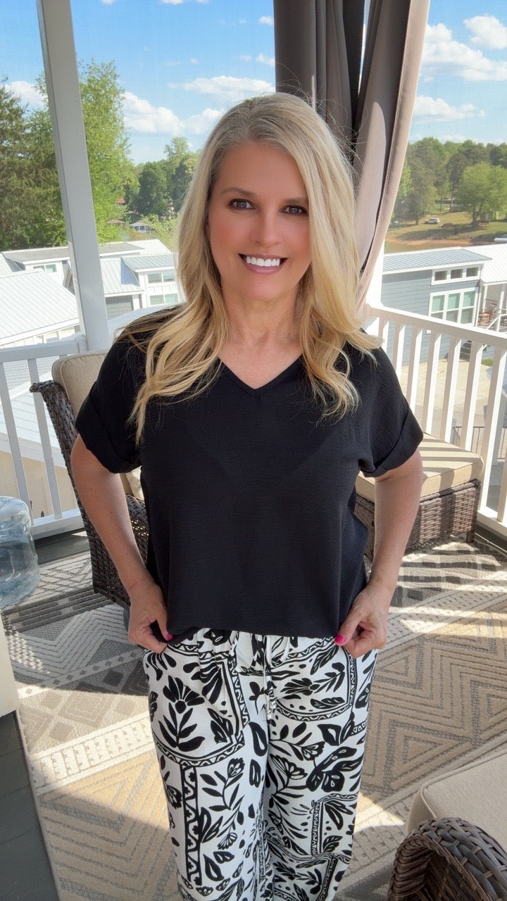Woman standing on a patio wearing a black top and patterned pants, smiling.
