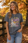 A woman wearing a dark gray graphic t-shirt with a church illustration and the text 'Be the Church When Service Is Over', paired with light blue jeans, standing outdoors in a park with autumn trees and fallen leaves.